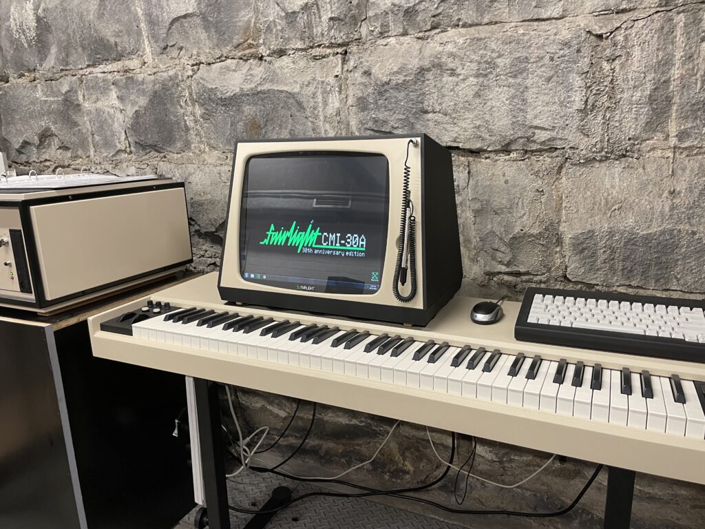 A Fairlight CMI synthesizer consisting of a monitor with green text on a black screen, a musical keyboard that is beige with white keys an on top of it, to the right is a vintage looking computer keyboard that is dark brown with beige keys.